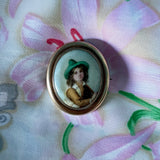 Antique brooch with a portrait of a  Hand Painted Victorian Tyrolean boy in a green hat on a fabric background.